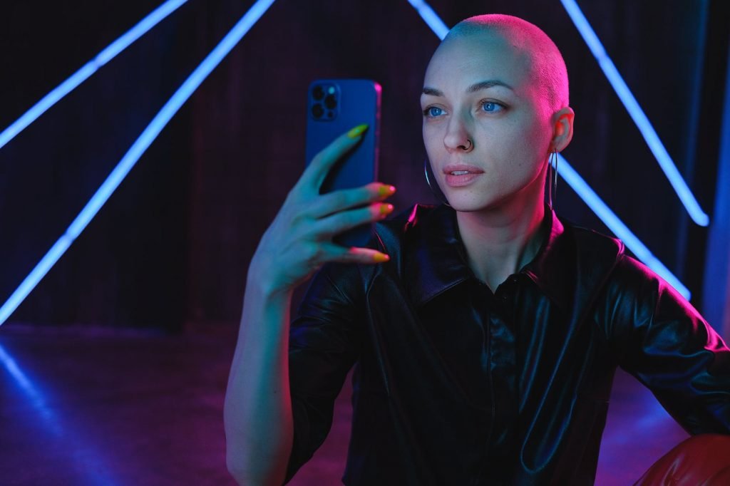 young bald woman using photos on smartphone in neon studio